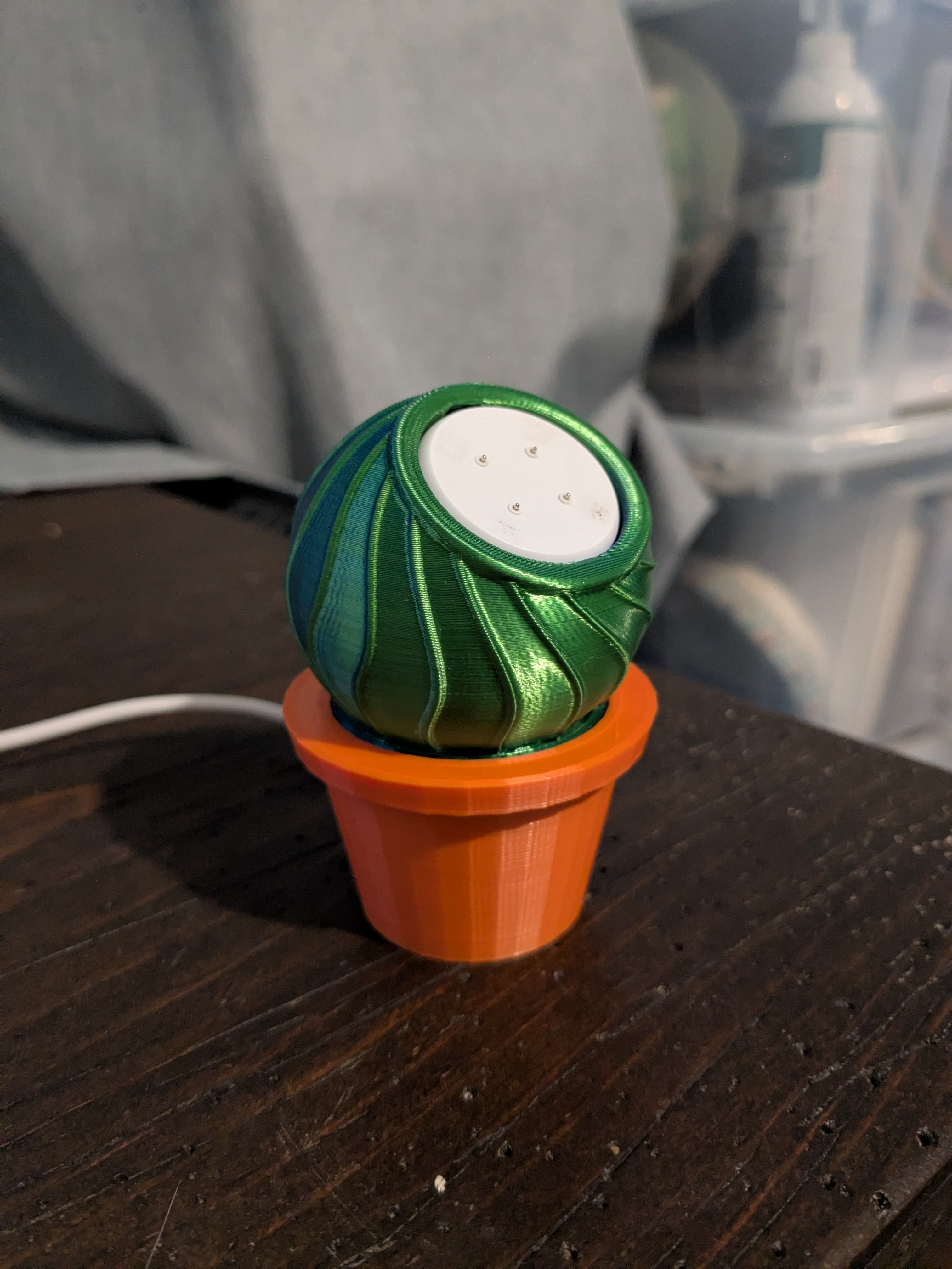 A small 3D printed sculpture shaped like a stylized cactus, with a swirled green and teal spherical top sitting inside a bright orange flower pot. A small power cord trails out, suggesting it lights up. It sits on a dark wooden tabletop with workshop supplies blurred in the background.