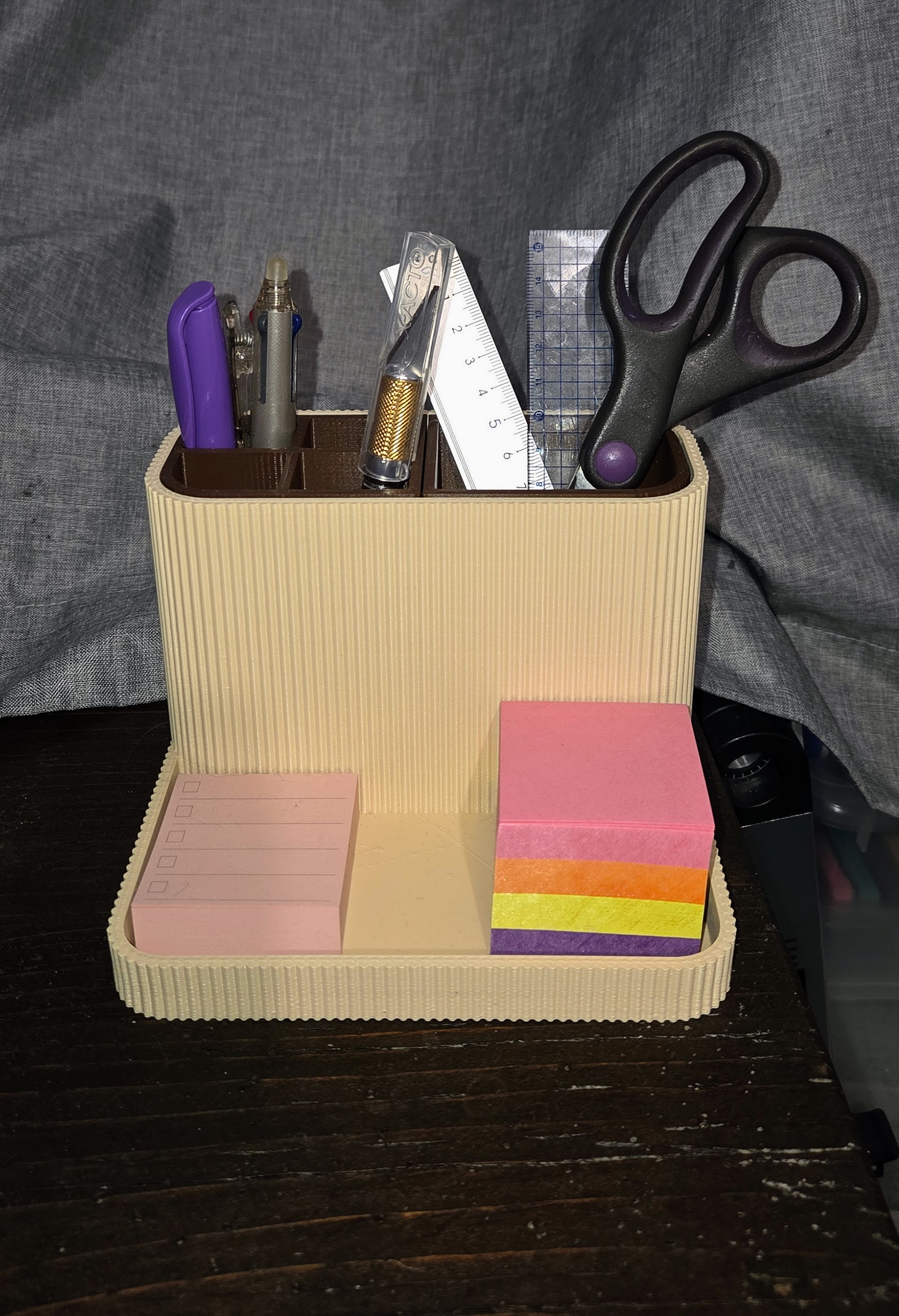 A 3D printed desk organizer with a creamy beige color and dark brown inserts. It holds office supplies including scissors, markers, rulers, pens, and small, rainbow-colored sticky note oads.