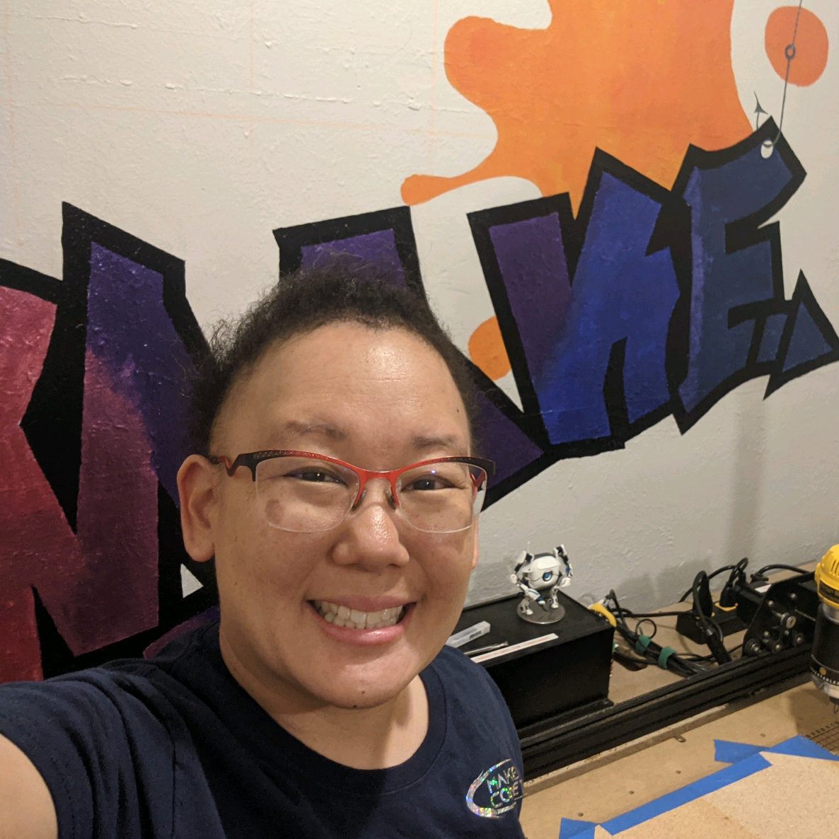 A warm portrait of Rei Cameron, founder of Maker Comet, smiling at the camera. She is Asian, with short black hair and wears thin metal-framed glasses and a dark navy t-shirt. Behind her, a white wall is painted with a colorful graffiti-style mural spelling 'MAKE' in stylized purple and blue block letters topped with an orange paint splash. To her right, a workbench holds small figurines and tools, hinting at the makerspace where she works.